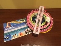 Photo of stacked colorful circular knitting looms with two purple needles and instruction booklet on wood surface against green wall.