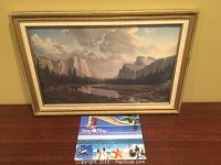 Framed oil painting showing wilderness landscape with mountains, river, trees and cloudy sky, set on wooden surface.