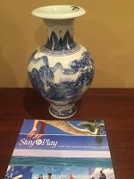 Full frontal view of vase displaying blue and white painted mountain and tree landscape motif. Size context given by brochure beneath.