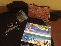 Photo of assorted stamped silver bracelets, a silver bangle with blue stone, silver chain necklaces, green jade ring, and a brown woven purse with gold chain strap on a black cloth background.
