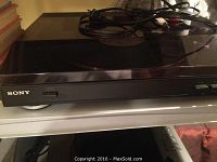Sony turntable record player with power and audio cables coiled on top, closed dust cover. Front left Sony logo visible.