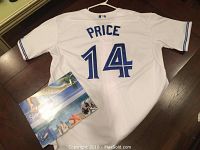 Back view of white Toronto Blue Jays jersey with 'PRICE' and number '14' in blue lettering, laid on floor with a catalog booklet nearby.