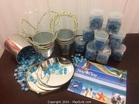 Multiple small plastic containers filled with various blue decorative beads, spilled beads, six small metal buckets with gold handles, and round reflective metal plates on a dark table.