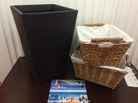 Photo showing the tall dark brown woven outdoor planter next to two stacked woven wicker baskets with white fabric liners and wooden handles.
