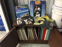 Wooden cabinet containing vinyl records and some displayed Elvis Presley albums on top, a Beatles shirt, and Stay & Play item