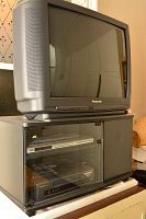 Panasonic 27-inch CRT TV on top of black cabinet with glass doors. Inside cabinet are Panasonic VHS player and JVC DVD player. Photo captures entire setup from side angle.