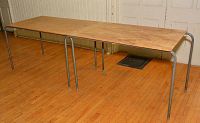 Long view of multiple stacked tables with plywood tops and metal legs in a room with wooden floors.