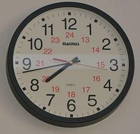 Magnus round wall clock with black frame and white clock face. Black hour and minute hands, red second hand, featuring both 12-hour and 24-hour numerals.