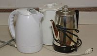 Photo showing three kitchen appliances: stainless steel coffee percolator, white plastic electric teapot, and white electric teapot which may be ceramic or metal, all with power cords.