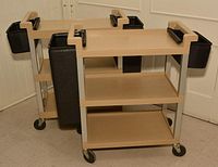 Two beige plastic 3-tier serving trolleys shown from the side, each with black plastic side containers for cleanup and caster wheels for mobility.