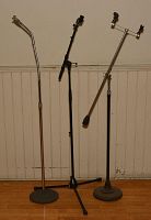 Photo showing three microphone stands against a wall: a silver stand with round base and gooseneck, a black tripod stand with adjustable height, and a chrome boom stand with round base and adjustable arm.