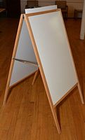 Photo showing wooden framed easel with whiteboard side visible, standing on a hardwood floor with chairs in background.