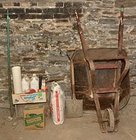 Mortar wheelbarrow, shovel, bags of Sheetrock and sweeping compound, and various small containers and bottles on the floor against a stone wall