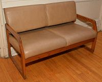 2-seater sofa with tan vinyl seat and back cushions framed in wood, angled armrests, placed on wood flooring.
