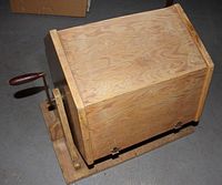 Side view of the wooden raffle ticket drum showing the crank handle and metal hinges.