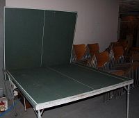 Fold-up ping-pong table with green top folded vertically and metal legs, partial view of stacked chairs in background.