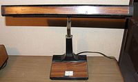 Front view of desk lamp showing rectangular lamp head with wood tone and black edges, on a black base with matching wood tone panel and two toggle switches.