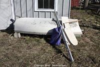 Hull flipped over resting on blocks; shows exterior top of hull, aluminum mast, blue sail bag, and rudder blades.
