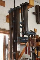 10 large F-type bar clamps hanging on a wooden rack in a workshop, showing handles and metal bars.