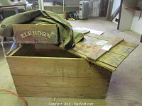 Large wooden crate with hinged wooden lid, visible markings and wear, situated indoors on wooden floor.