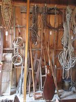 Wide view of multiple long wooden boat parts hanging and leaning including oars, rudder, and ropes in a wooden shed.