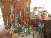 Overall view of garden tools and supplies arranged against wooden wall with various rakes, shovels, pots, bird feeders, and other items