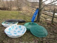 Wide outdoor scene showing two kiddie pools with different prints, two toboggans in black and blue, green Little Tikes turtle sand box with cover, and wooden rope swing hanging from tree.