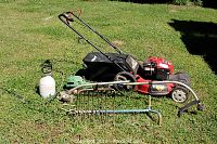Full view of all lot items laid out on grass: red Troy-Bilt lawn mower, green John Deere edger and weed eater attachments, metal pitchfork head, metal garden claw tool, white plastic liquid container.