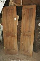 Two wood panel doors leaning against a structure, showing overall height and panel design.