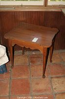 Full view of the smaller wooden side table showing the scalloped edges and tapered legs with caster wheel on an orange tile floor.