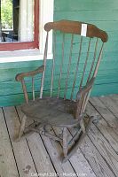 Full view of wood rocking chair showing contoured seat, spindle backrest, armrests, and curved rockers on a wooden porch floor.