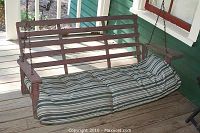 Brown wooden porch swing with horizontal slats and striped cushion, showing right arm detached.