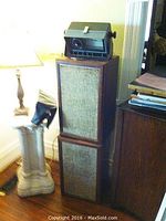 Two stacked Scott speakers with wooden casing and fabric grill, placed on the floor next to a lamp stand and other furniture.