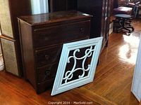 Photo showing wood chest of drawers with three drawers, dark finish, and detached handle on top left drawer with white decorative wood art panel leaning against it.