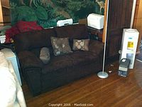 Brown fabric sofa with cushions, including two decorative pillows with circular patterns and a bolster pillow. Sofa is positioned on a wooden floor with a painted wall backdrop.