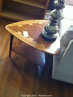 View of wooden side table showing triangular shape, wood grain and inlaid wood pattern, with lamp on top next to sofa.