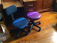 Photo showing two rolling chairs (blue seat and back; purple circular seat without backrest) in front of vintage wooden desk with drawers and opening top.