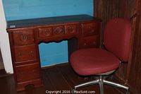 Photo showing wooden desk with multiple drawers and maroon office chair with metal rolling base placed beside it.