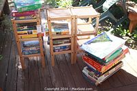 Overall view of collection showing the stack of classic games and puzzles alongside three wooden storage shelving units arranged outdoors on a wooden deck.