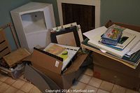 Photo showing white display shelf, several cardboard boxes, plastic bin with art supplies including papers and sketch pads.
