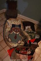 Photo of wooden teddy bear children's chair, two baby cradles with bedding, woven basket, dolls and toys grouped on tiled floor against a wall