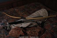 Photo showing two metal baseball bats, two brown catcher's mitts, a wooden bow, and a gray fabric case on a bed with patterned blanket