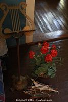 Photo of a metal ornate lyre-shaped music stand with platform, next to a pot of artificial red flowers and two small model sailboats on wooden floor.