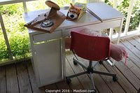 White child's desk with drawers, red swivel chair positioned in front, folded white chair leaning on the side, wooden puzzle, stuffed animal toy, and pink rolled item on top of desk. Outdoor setting with natural cypress or trees in background.