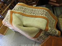 Synthetic fibre area rug folded and draped on wooden furniture showing backside and beige geometric pattern with brown trim.