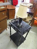 Black faux wood accent table with lower shelf shown with a black lamp with white lampshade on top, lamp cord visible.