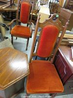 Two wood dining chairs (one armchair and one side chair) with rust velvet upholstery, showing wear and scratches, situated indoors among other furniture.