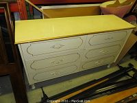Photo showing front view of 6 drawer cream-colored dresser with decorative trim and ornate handles, visible wear and some damage.
