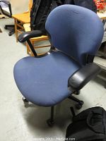 Front angle of blue office chair showing fabric seat, backrest, and black plastic armrests with castor wheels.