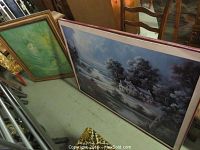 Two framed prints leaning against a cabinet and wall: one with a scenic rural landscape and the other a green-tinted portrait-style image.